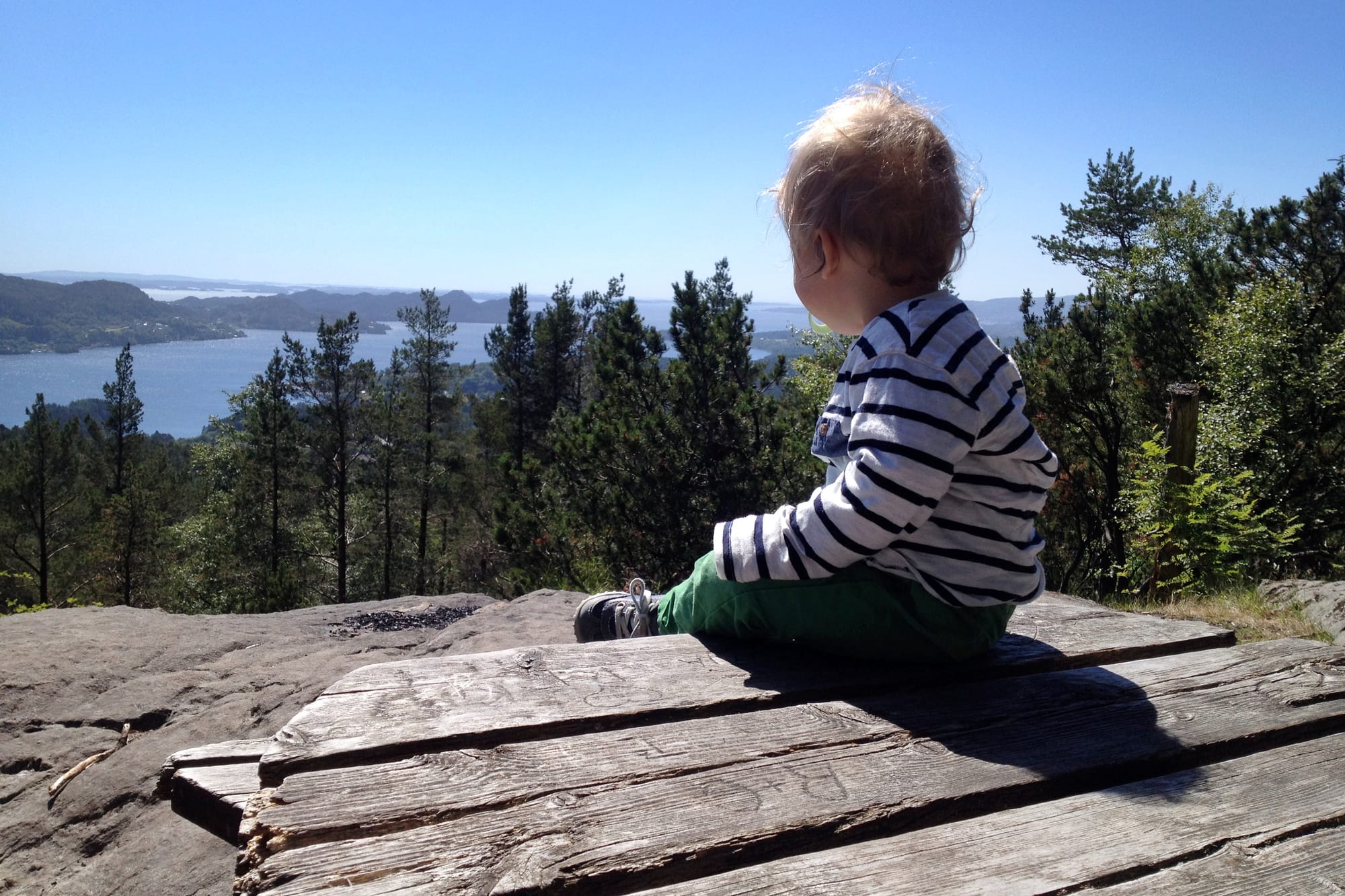Et barn sitter på et trebord og ser utover en utsikt med innsjø, skog og fjell under en klar blå himmel.