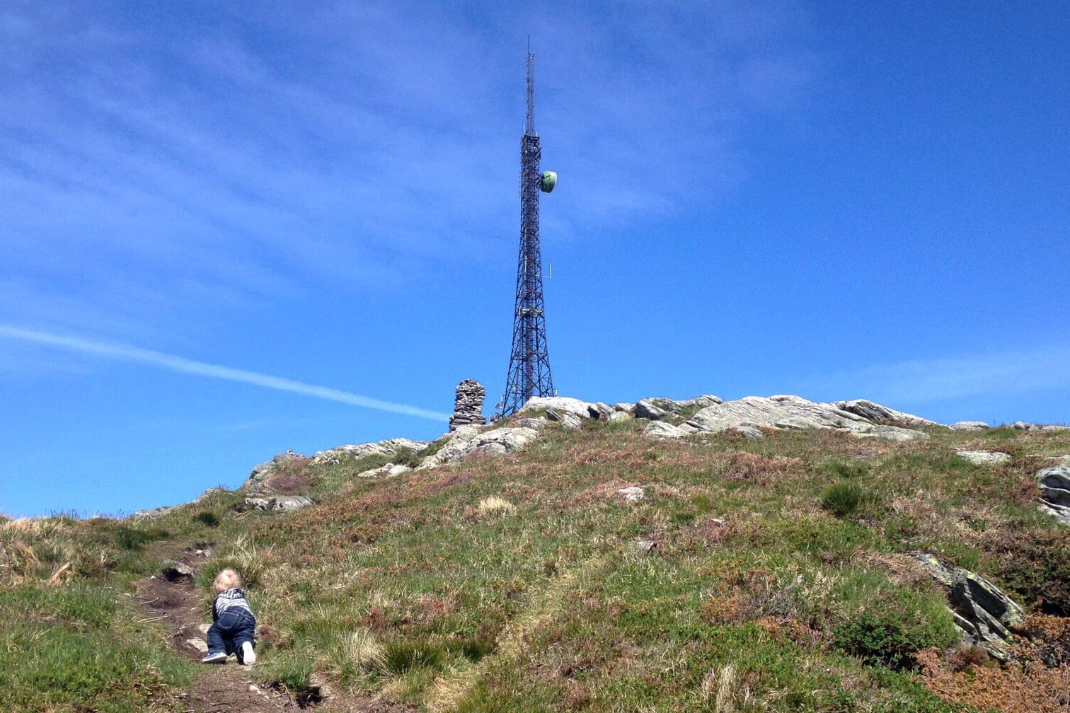 Et barn klatrer opp en sti mot en radiomast på en gresskledd fjelltopp, med en steinvarde ved siden av, under en klar blå himmel.