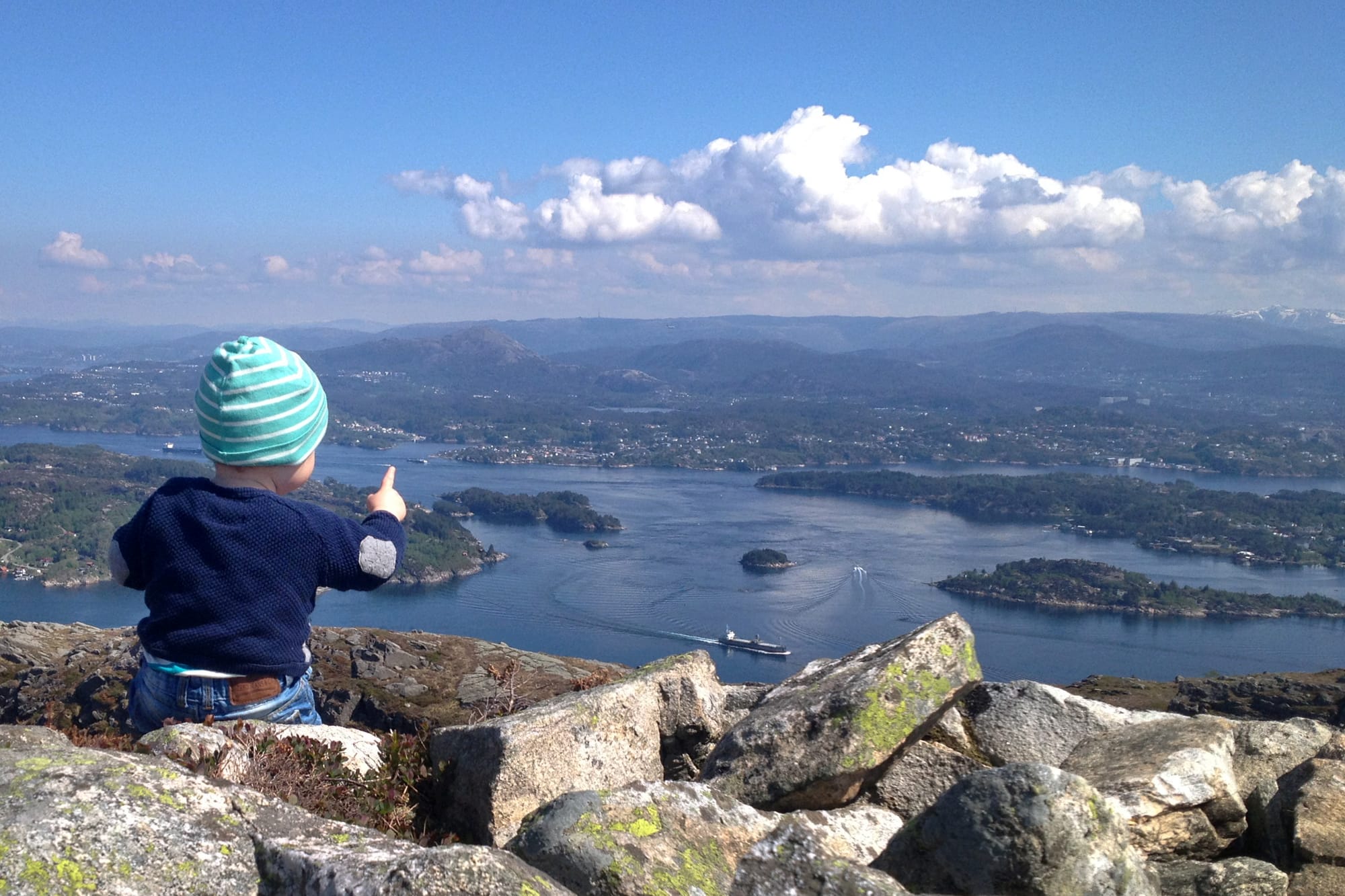 Et barn med blåstripet lue sitter på en fjellkant og peker utover fjorder og øyer under en klar blå himmel med spredte skyer.