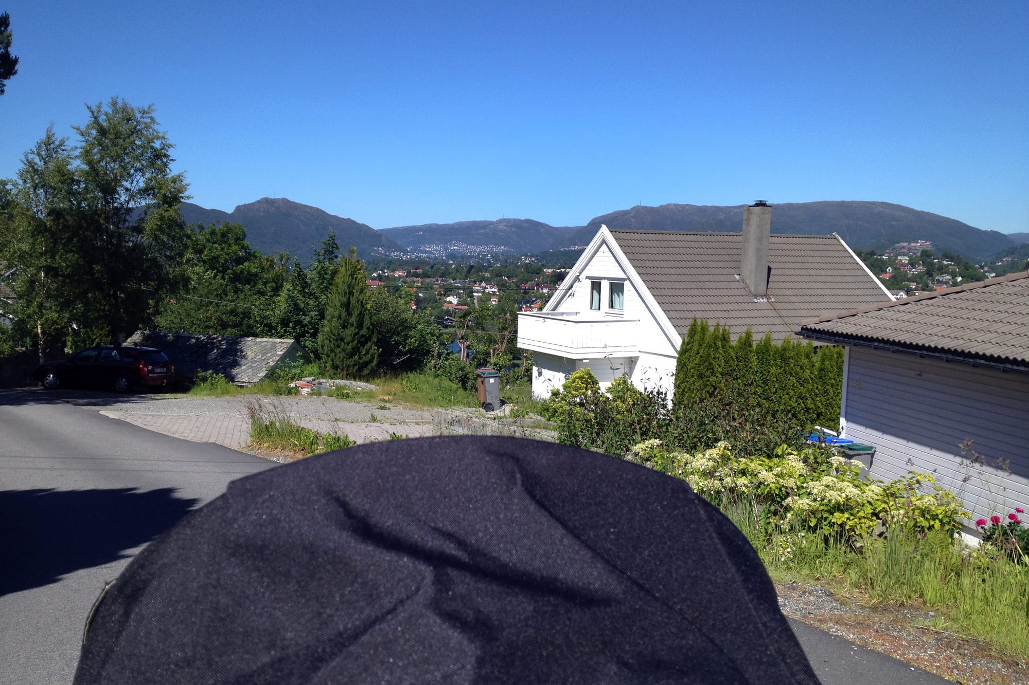 Utsikt fra en barnevogn mot Byfjellene, med hvite hus, trær og en blå himmel i forgrunnen. Byen skimtes i det fjerne.