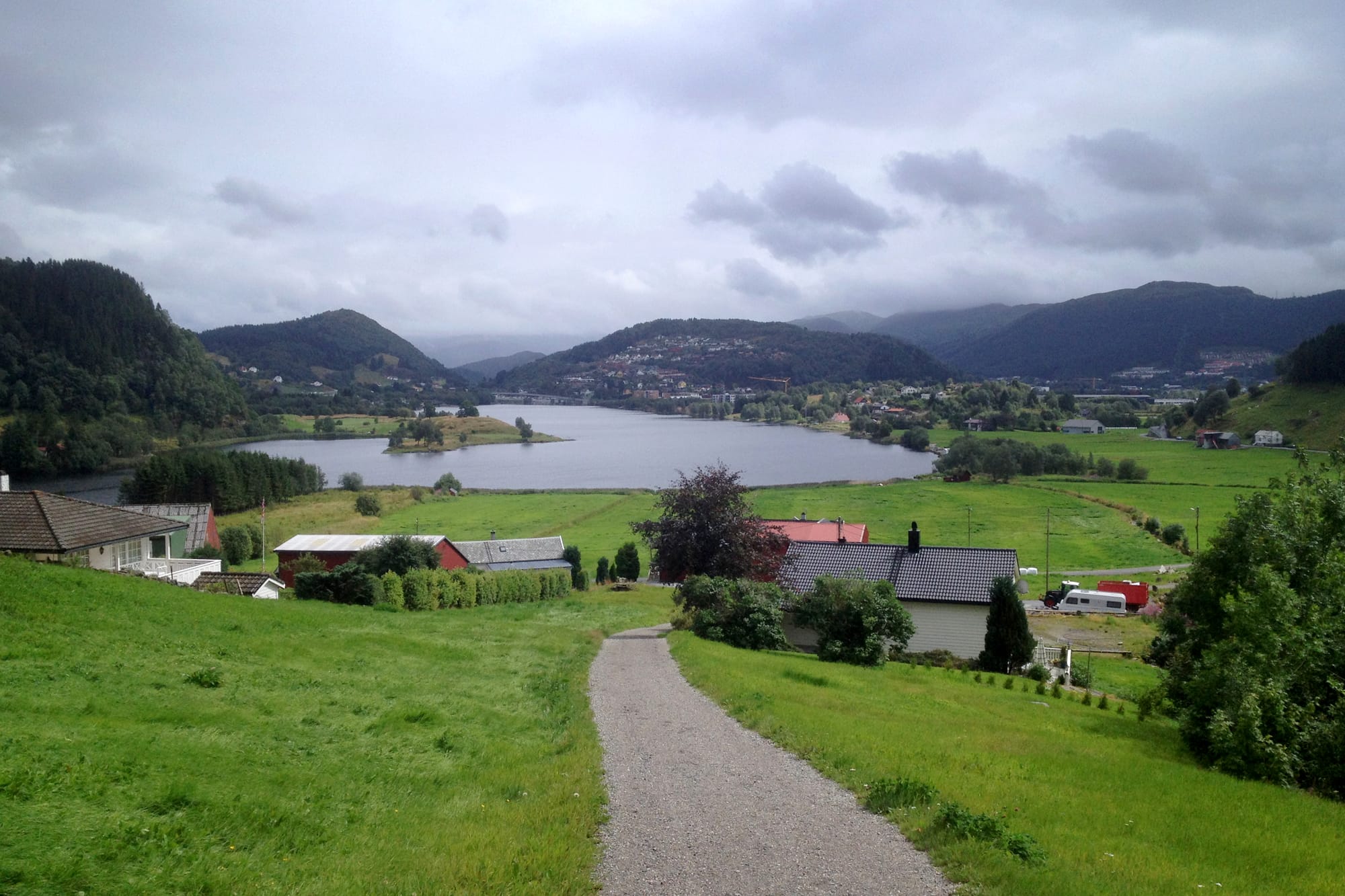 En grusvei leder ned til en innsjø omgitt av grønne marker, hus og skogkledde åser under en overskyet himmel.