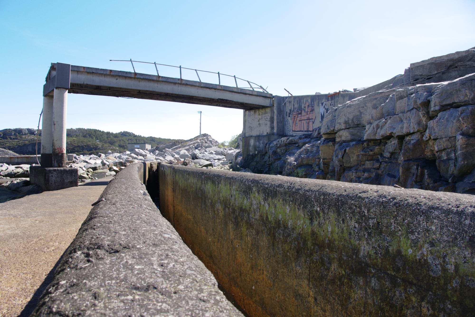 En rusten betongbro står over en smal renne omgitt av stein og betong. Graffiti dekorerer en vegg, under en klar blå himmel.