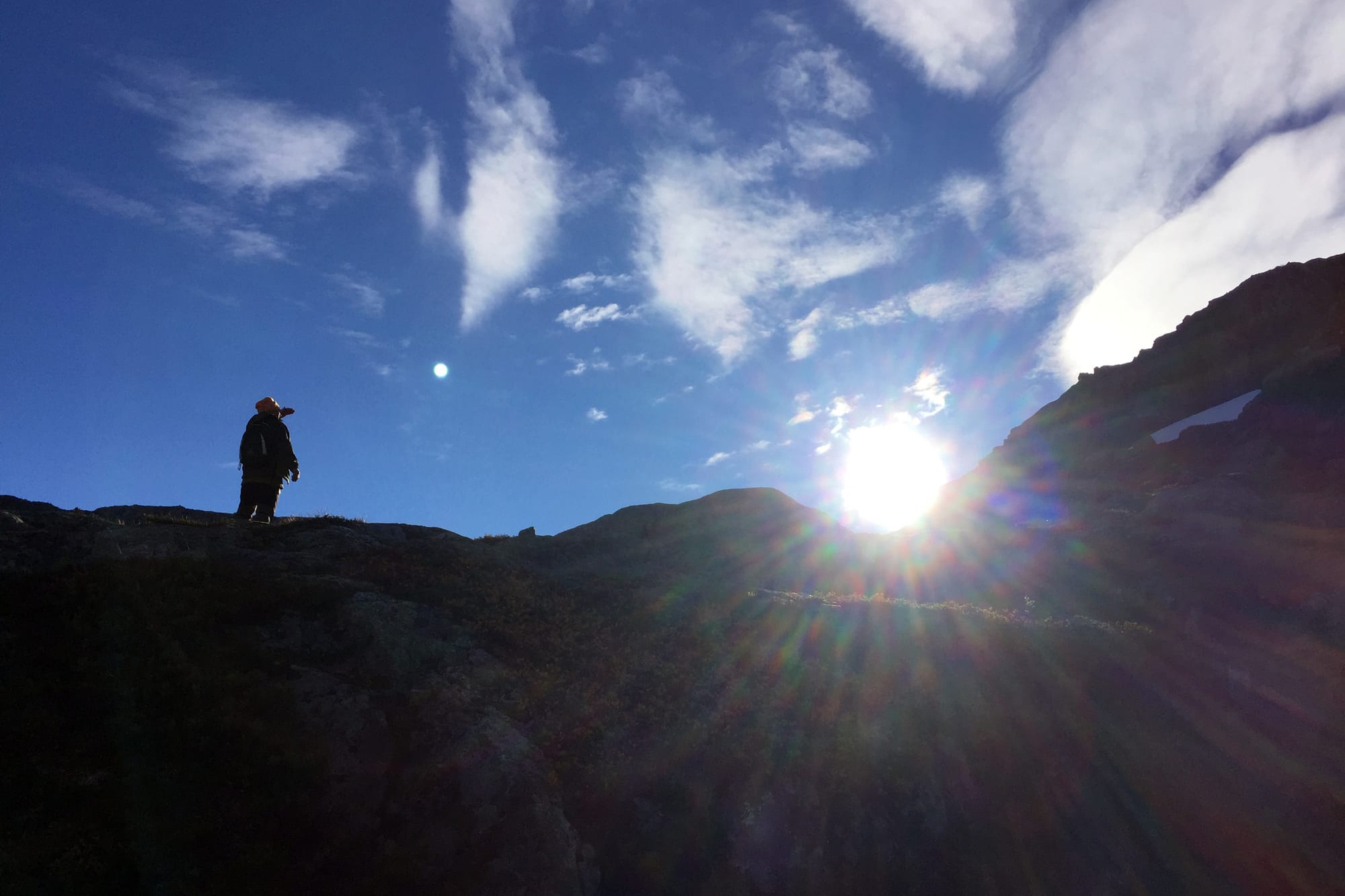 En turgåer står på en fjellhylle, badet i strålende sollys. Blå himmel og skyer danner en dramatisk ramme rundt opplevelsen.