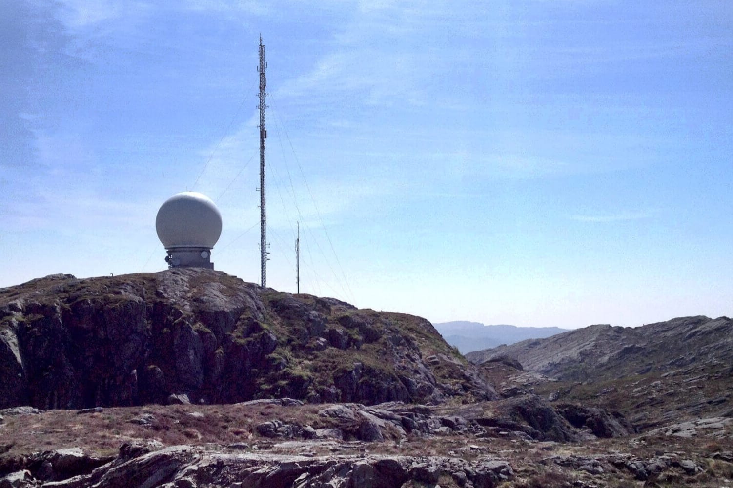 En radarstasjon med en hvit kuppel og høye antenner står på en fjellrygg omgitt av lyng. Klar blå himmel i bakgrunnen.