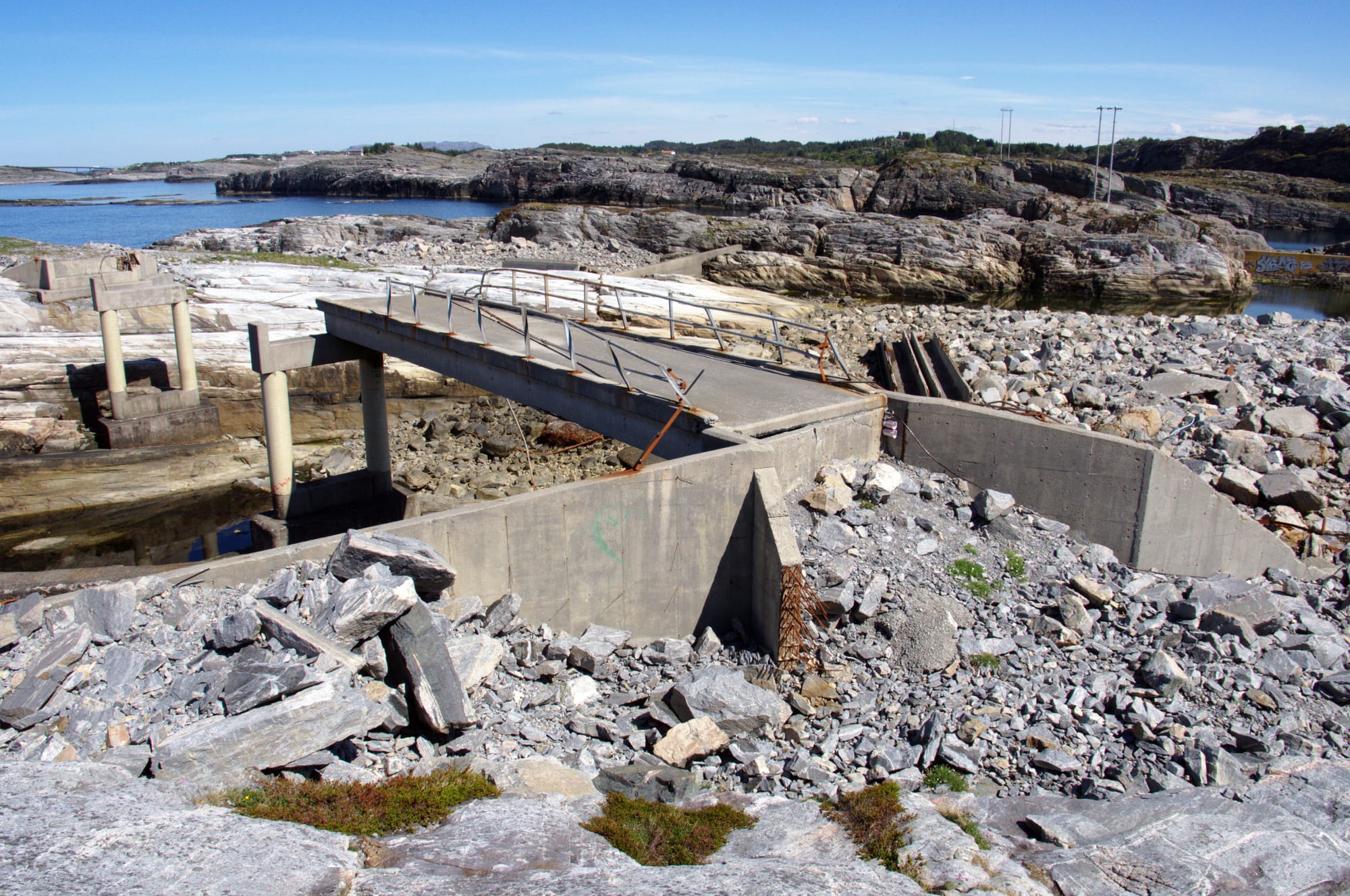 Ødelagte rester av en betongbro over en steinfylt kløft, omgitt av kystlandskap og vann under en klar blå himmel.