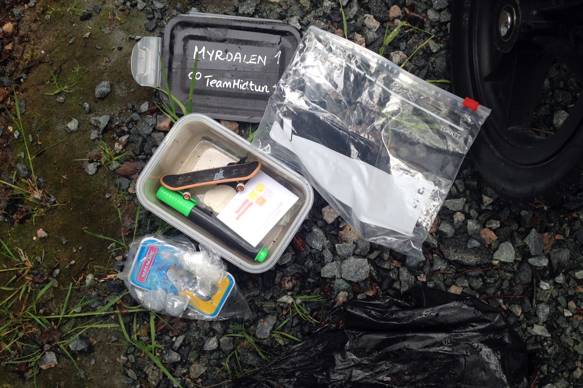 En geocache med plastbokser, zip-poser og små gjenstander, inkludert en miniatyr-skateboard, lagt ut på grus og mose.