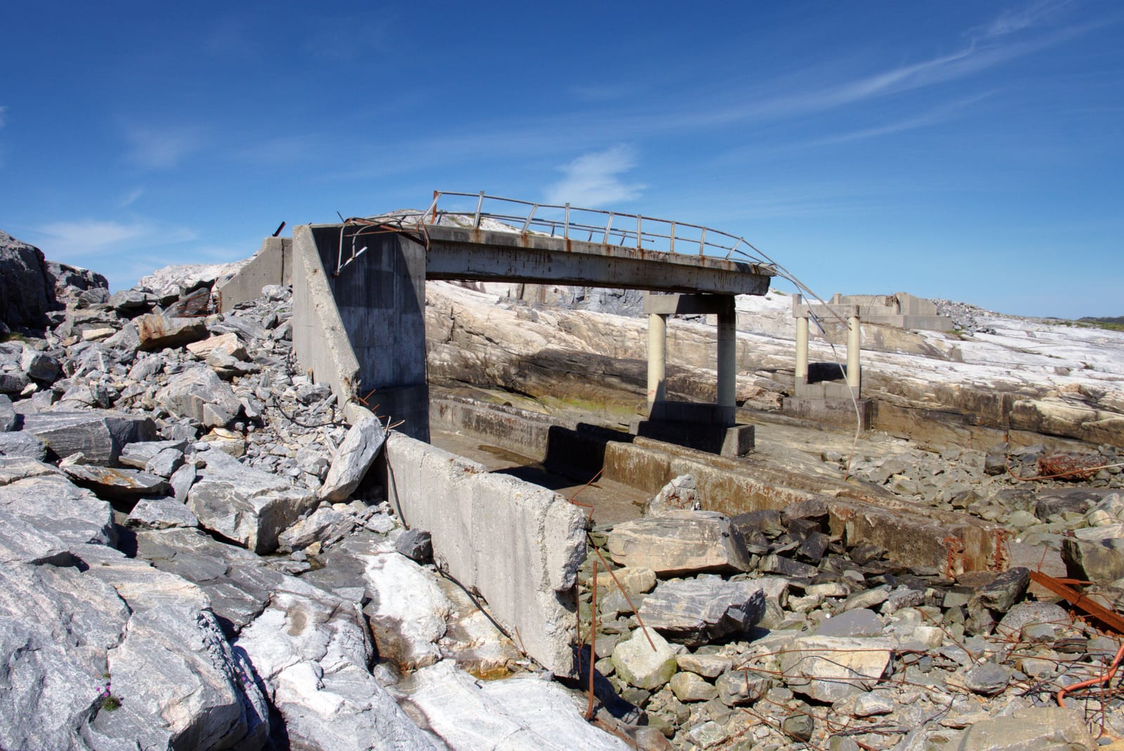 En ødelagt betongbro står delvis over en steinete kløft, omgitt av knuste steiner og rustne armeringsjern under blå himmel.