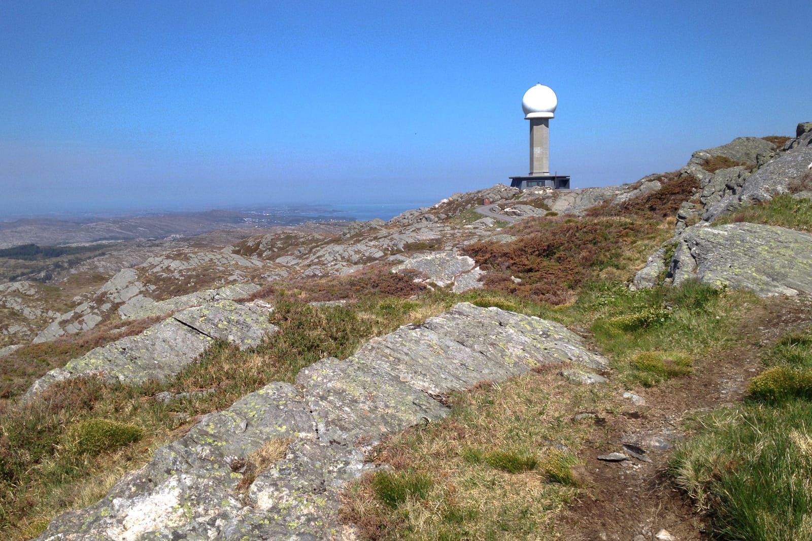 Fjellandskap med lyng, steiner og stier. På toppen står en radarstasjon med hvit kuppel, omgitt av blå himmel og kysttutsikt.