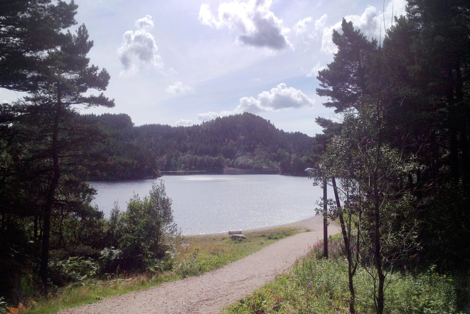 En grusvei fører til en innsjø omgitt av skogkledde åser. Solen skinner på delvis skyet himmel over et idyllisk landskap.